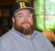 Headshot of John Dewees in a checked shirt, glasses, and baseball cap, in front of a window