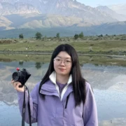 Headshot of Kexin Zhang, in glasses and a purple jacket, holding a camera in one hand, in front of a lake and mountains